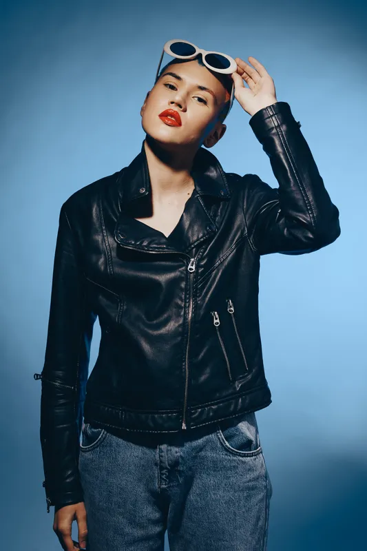 Stylish woman wearing black leather jacket and sunglasses poses against blue background, showcasing modern fashion, attitude, and casual street style.