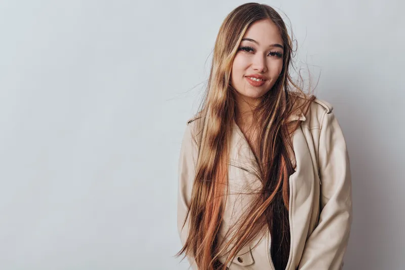 Young woman with long hair smiling in beige jacket standing against plain background casual fashion style confidence and beauty portrait indoors