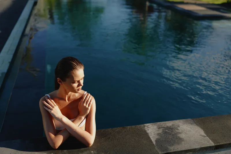 Relaxed woman by a sunlit pool edge, resting on the concrete, enjoying a peaceful moment by the water. Outdoor lifestyle scene emphasizing leisure, warmth, and self care with soft light and tranquil
