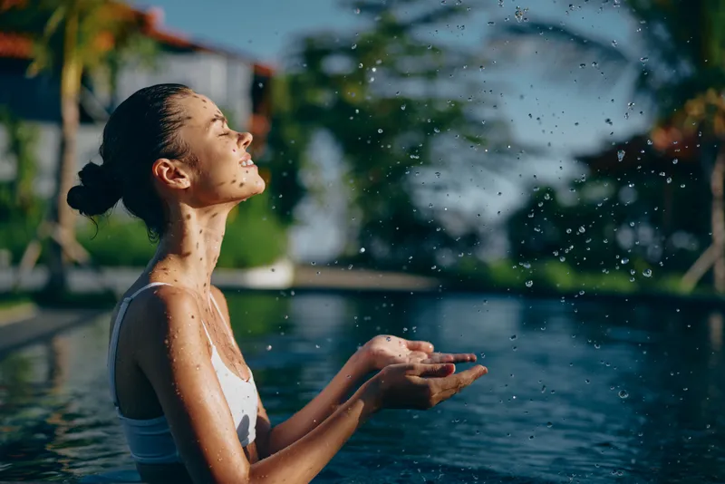 An outdoor poolside moment shows a woman facing the sun, water droplets rising as she cups her hands. A clear lifestyle concept emphasizing relaxation, wellness, and leisure in a bright, warm setting.