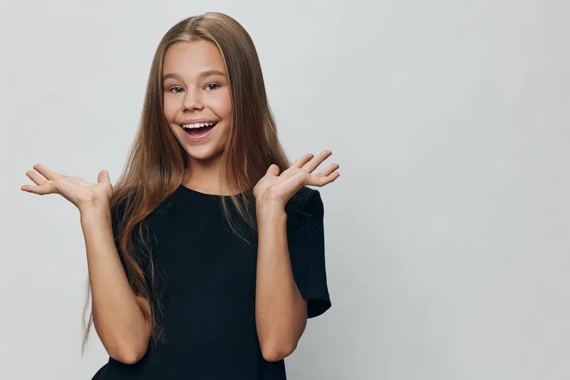 Girl smiling and showing excitement with open hands and long hair in casual black shirt on grey background. Happy child with expressive face and bright eyes in studio portrait.