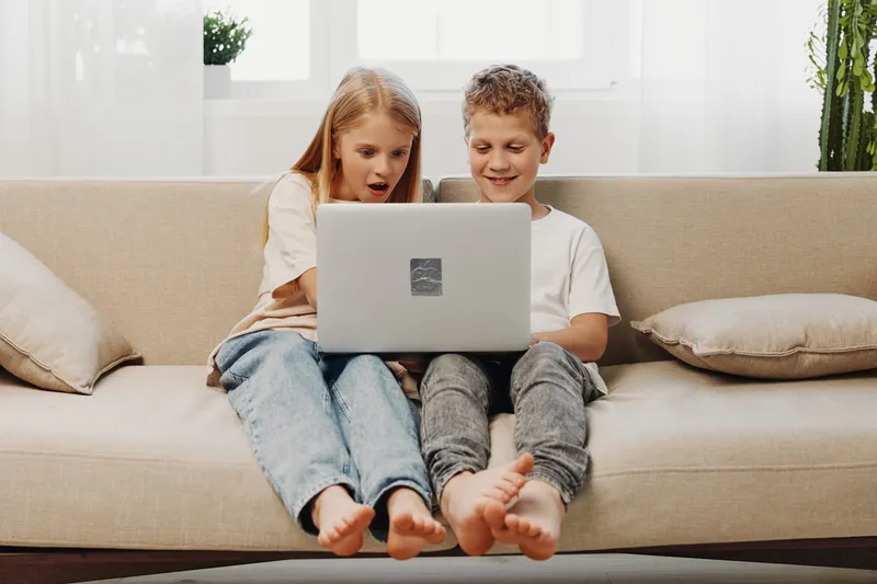 Two children sit barefoot on a sofa using a laptop together, showing surprise and joy. The bright living room setting highlights casual home entertainment and shared digital experience.