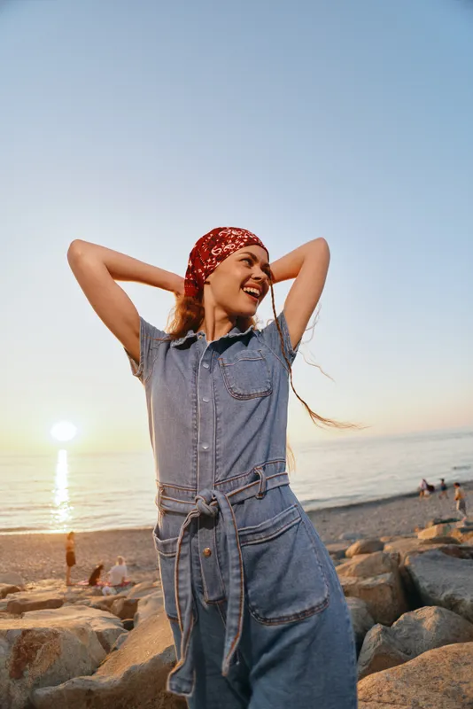 Smiling woman in denim dress with red headscarf enjoying sunset by the rocky beach, feeling relaxed and happy outdoors.