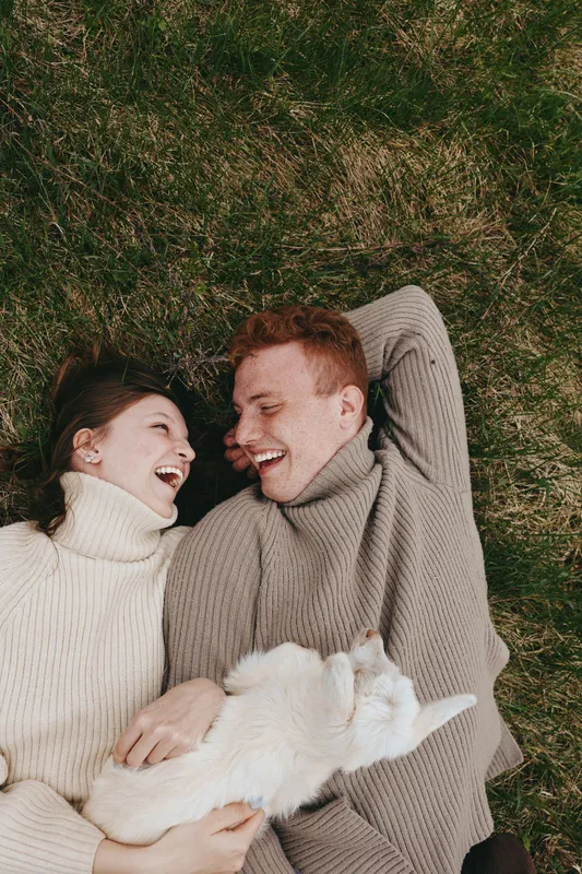 Happy couple lying on grass in sweaters, a fluffy cat between them, cozy outdoor lifestyle moment with warm tones, intimate candid scene, relaxed mood, casual affection, nature backdrop, smiling faces