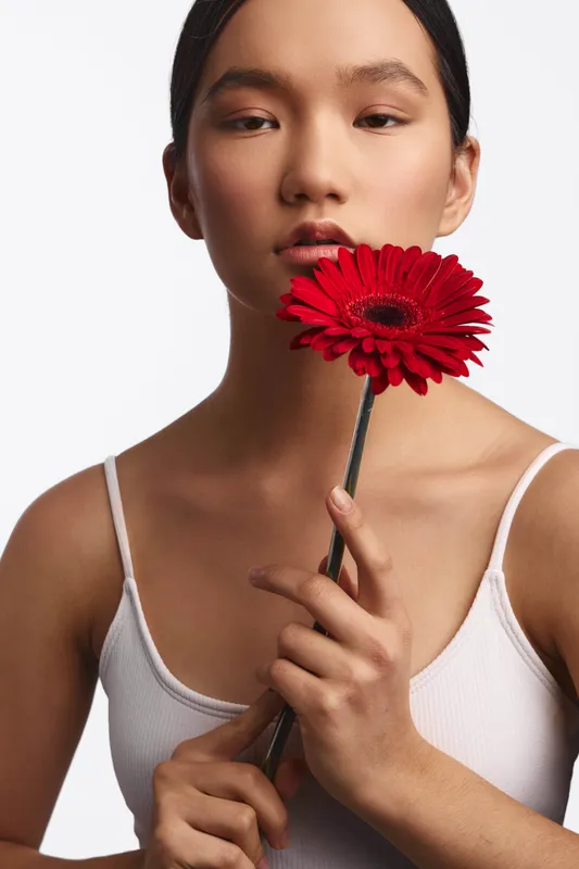 asian woman with a flower close to her face, wearing a white tank top, minimalist studio portrait highlighting delicate features and a contemplative mood.