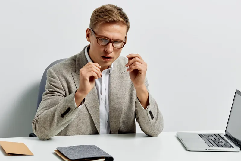 Professional man in formal attire and glasses sitting at office desk with laptop, looking surprised or confused while working on computer. Interior background, modern workspace, business lifestyle con