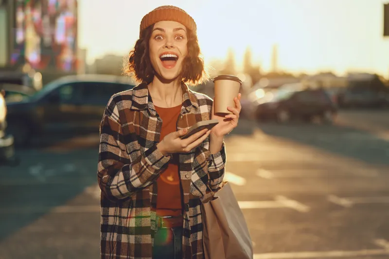 Woman holding coffee and phone on urban street with a bright smile in warm sunlight, candid moment of happiness and authenticity, golden hour glow and mindful living, emotional storytelling.