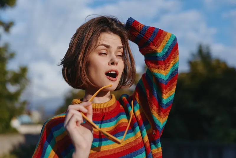 Woman portrait in a rainbow sweater holding sunglasses outdoors with expressive expression and casual pose in warm sunlight, blurred background for lifestyle, fashion and candid portrait use