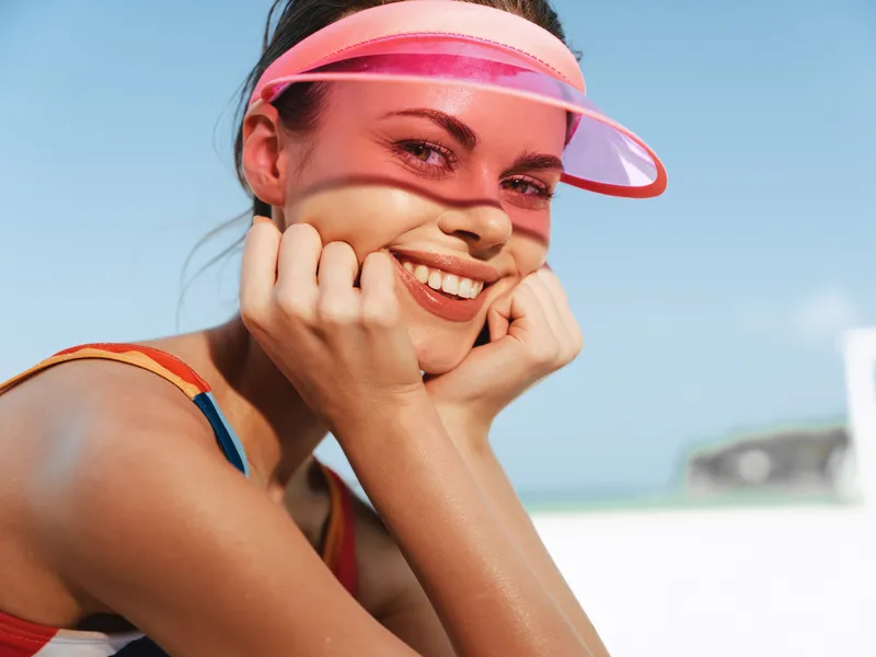 Close-up of a smiling woman at the beach wearing a pink visor, sunlit skin, and colorful swimsuit straps, capturing carefree summer vibes, warm air, and outdoor happiness.