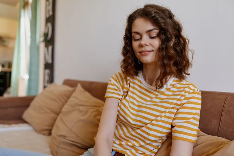 Casual woman indoors, relaxed on a couch, striped yellow and white tee, soft natural light, warm home mood, candid lifestyle moment, relaxed posture, gentle ambience, simple comfort and serenity