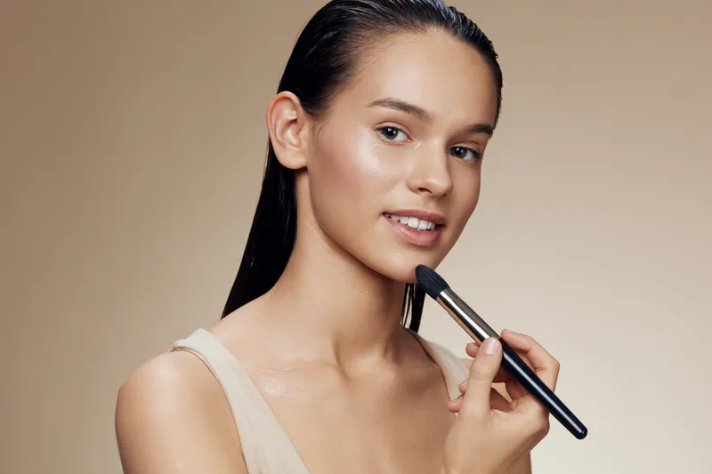 Portrait of a young woman applying makeup with a brush, showcasing a natural beauty look on a soft beige background, highlights a fresh, elegant, minimalist aesthetic