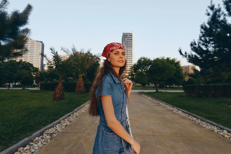 young woman wearing denim dress and red bandana walking in urban park during golden hour with modern buildings and green trees in background