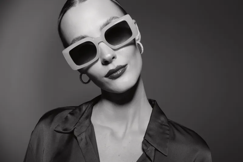 Stylish white woman with pale skin wearing large sunglasses and earrings poses confidently in black shirt in studio lighting.