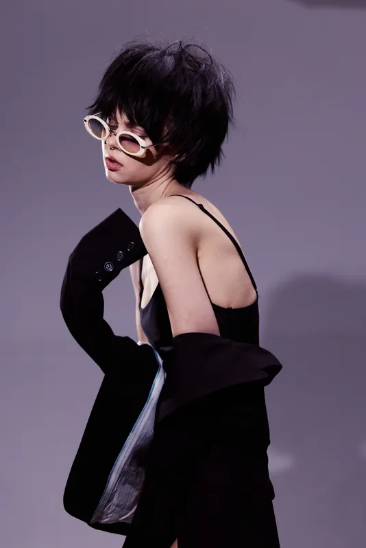 Fashionable woman with short hair wearing sunglasses and a black outfit posing on a purple background.