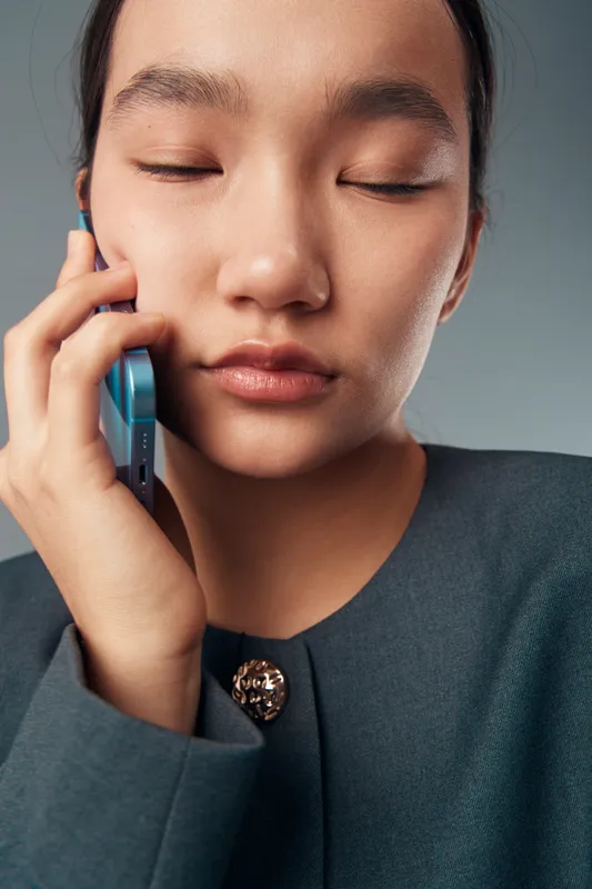 Woman with closed eyes holding phone to ear in closeup portrait. Concentrated young female talking on mobile smartphone with calm expression wearing dark jacket with decorative button.