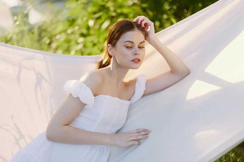 Young woman in white dress relaxing in hammock surrounded by greenery, expressing calmness and serenity in a vibrant summer setting