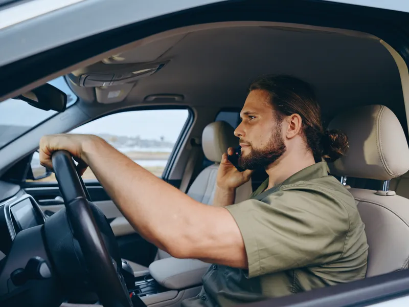 Portrait of a focused man driving a car while talking on a mobile phone, gripping the steering wheel, and maintaining alert posture during daytime commute.