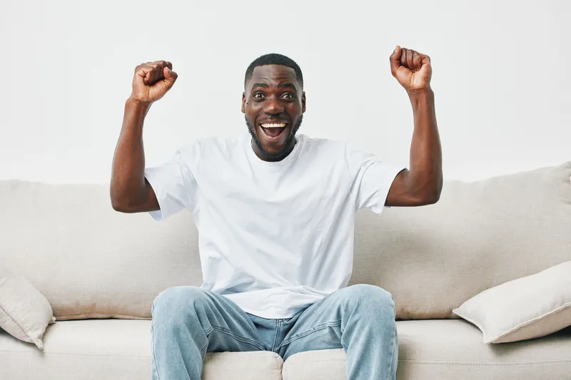 Happy young man celebrating on a cozy sofa, wearing a casual white t shirt and jeans, expressing joy and excitement in a bright, minimalistic living room