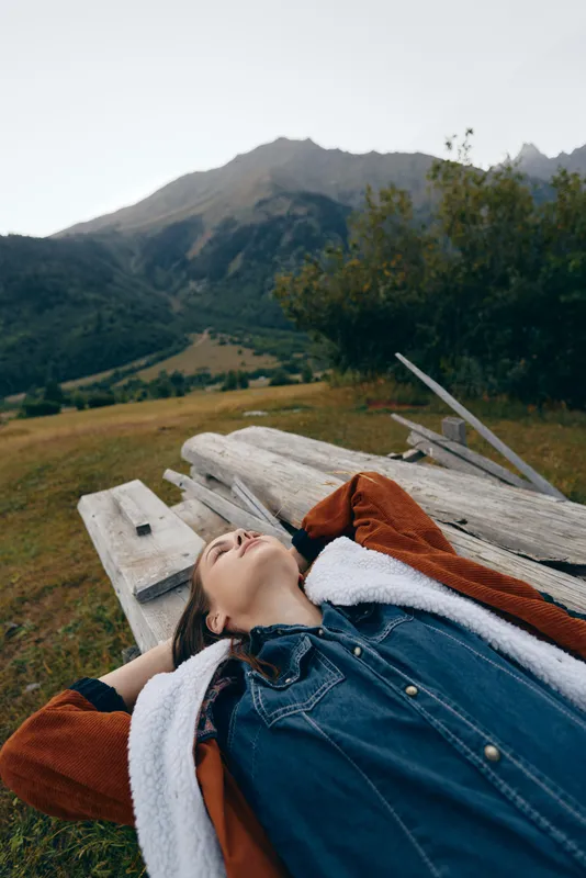 Woman relaxed on wooden bench outdoors in nature, portrait of casual traveler lying back in denim and shearling jacket on meadow with mountain backdrop and peaceful expression