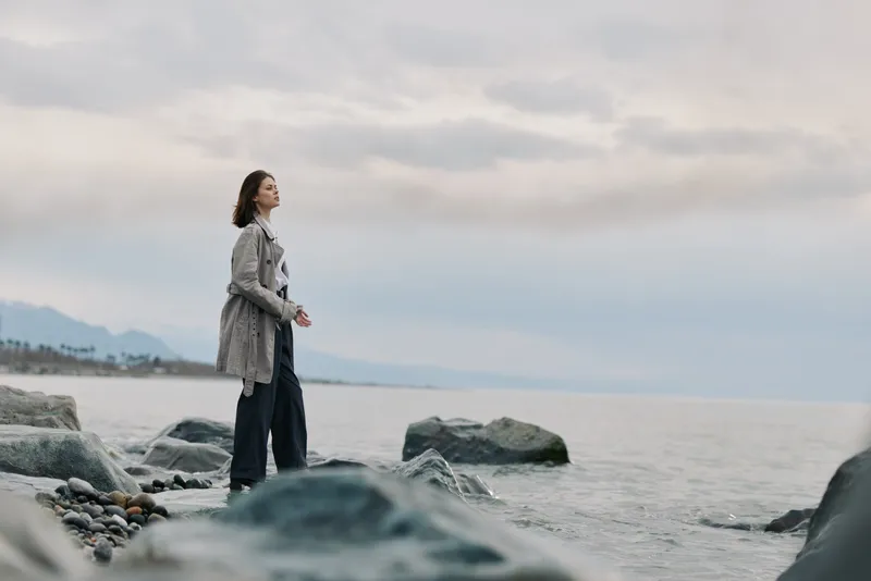 Young woman stands on rocky shore looking at calm sea under cloudy sky. Concept of solitude, nature, and peaceful moment by the water.