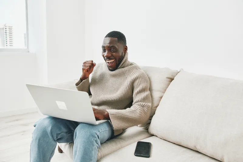 Happy young man celebrating success while working on a laptop at home, wearing a cozy sweater and relaxed jeans, showcasing a positive remote working vibe