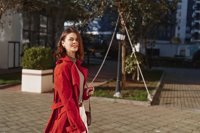 Young woman with brunette hair wearing a red trench coat walks confidently in an urban park during autumn, enjoying the warm daylight and fresh air.