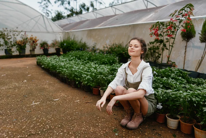 Young woman enjoying gardening in a greenhouse, surrounded by lush green plants in pots, wearing rustic clothing, depicting joy and connection with nature