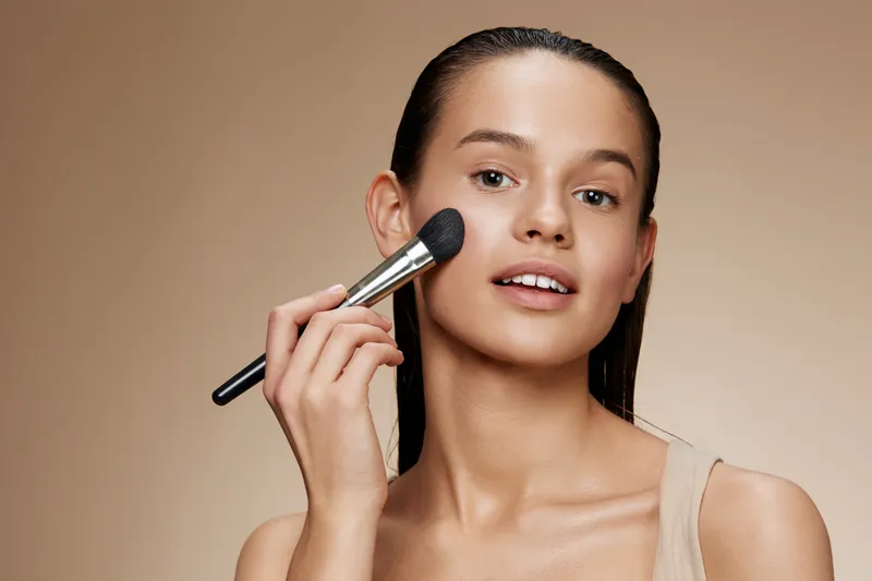 Young woman applying makeup with a blush brush, showcasing natural beauty and confidence against a warm beige background