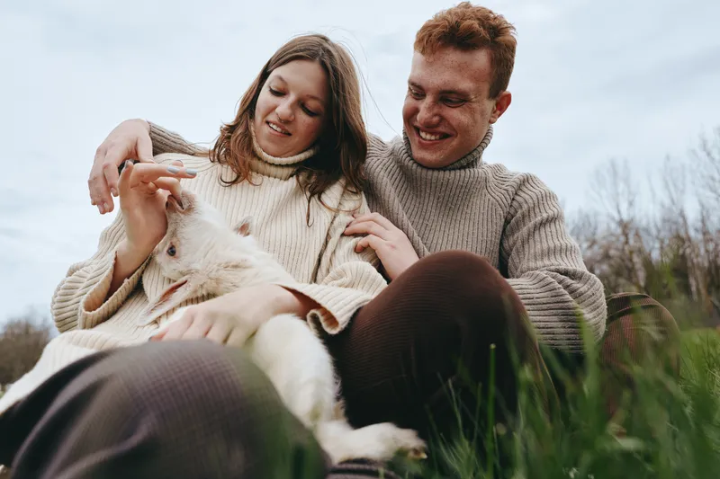 Cheerful couple outdoors with a playful goat kid nearby, cozy sweaters, relaxed pose, sunlit meadow, natural colors, warm family vibe, candid connection, rural lifestyle