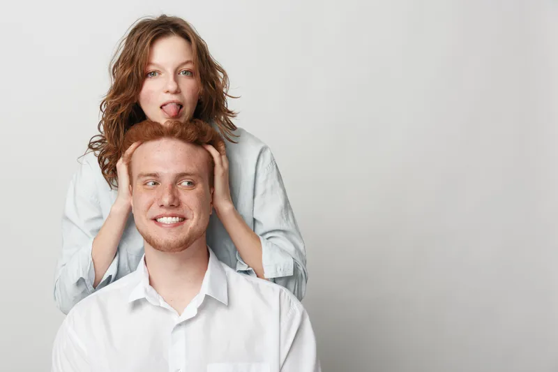 Young playful couple with red hair and casual clothes posing on grey background. Woman stands behind man holding his head and making funny face. Concept of friendship and fun moments.