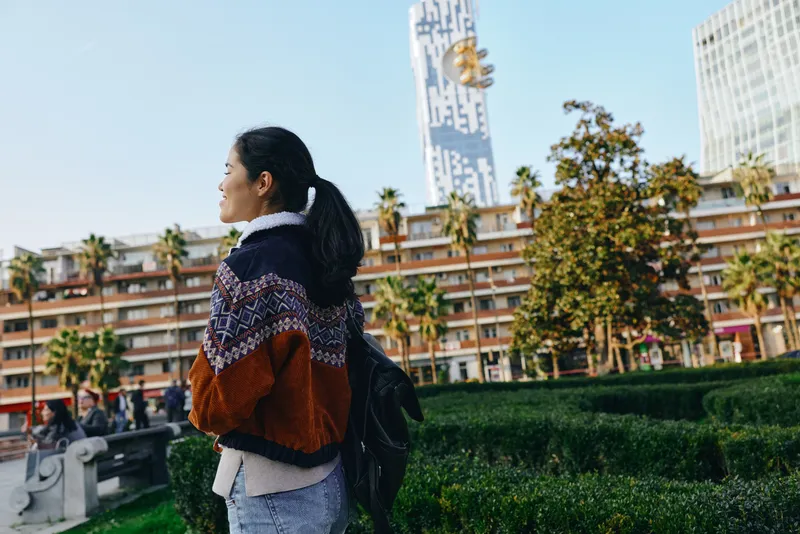 Woman with a backpack and chunky sweater walks along a sunny city street, surrounded by greenery and modern architecture, capturing outdoor exploration and everyday urban life.