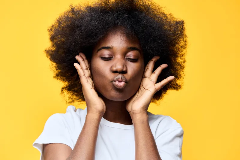 Happy young woman with voluminous afro hairstyle making playful face against bright yellow background, expressing joy and confidence