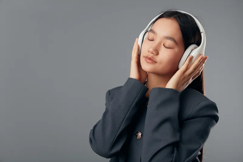 Young woman wearing headphones and closed eyes listening to music or podcast with peaceful expression on gray background in studio for relaxation and sound enjoyment.