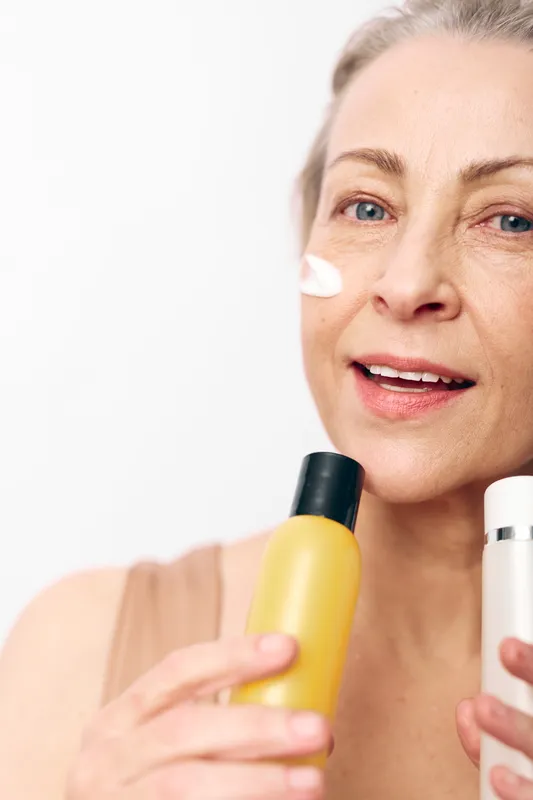 Mature woman with natural skin applies cream on face holding skincare bottles in hands. Bright studio background highlights wellness, beauty, self-care, and healthy lifestyle concept.