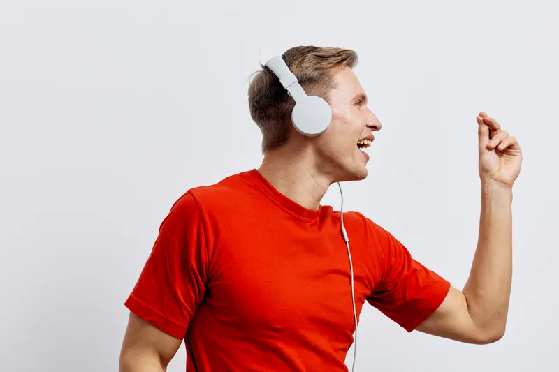 Man wearing red t-shirt enjoying listening to music with white headphones, smiling and dancing joyfully in motion isolated on plain white background studio portrait. People lifestyle concept