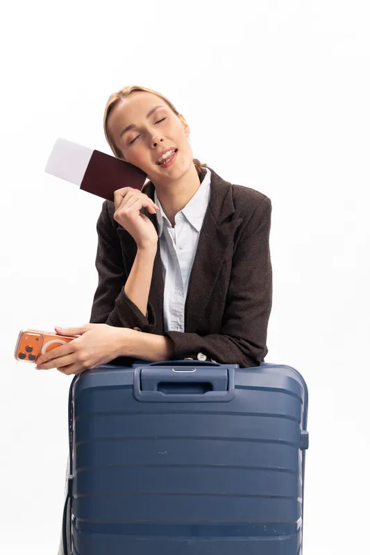 Tired young woman in business attire holds passport and boarding pass, leaning on blue suitcase. Travel concept showing exhaustion before a trip in studio with white background and casual mood.