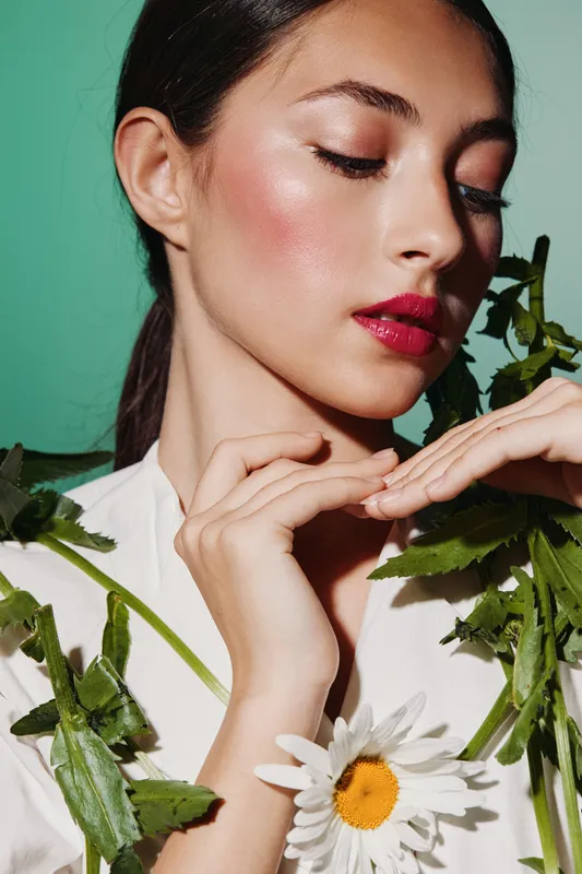 Close-up portrait of a young woman with glossy makeup framed by fresh green leaves and a daisy flower, on a bright green background. Artistic beauty fashion concept.