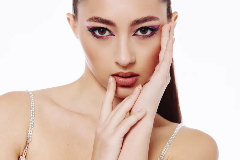 Closeup of a fashionable young woman with striking bright eye makeup and glossy lips posing with hands framing her face against a white background. Concept of beauty, fashion, and creativity.