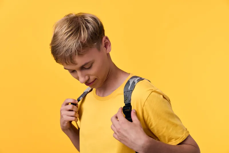Young student in a yellow t shirt adjusts backpack straps against a vibrant backdrop, ready for class, showcasing focus, preparation and daily school routine.