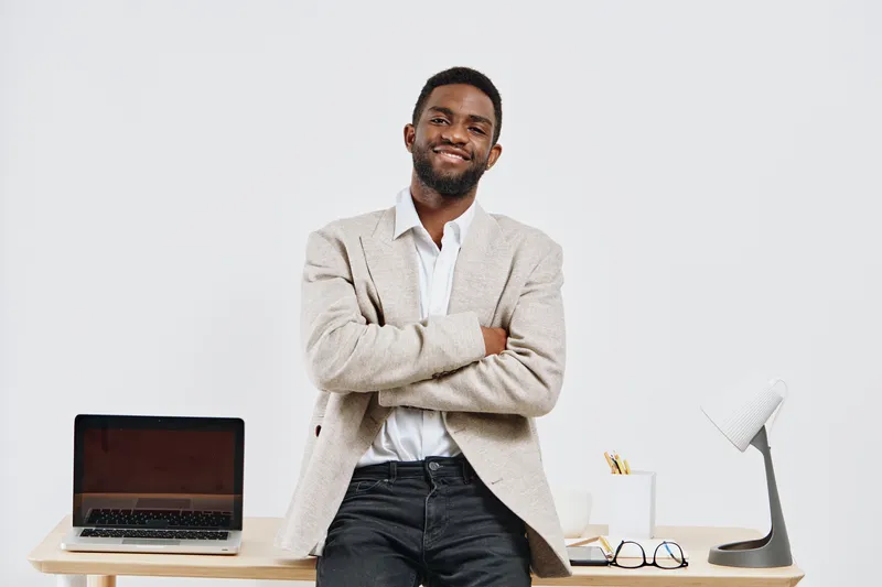 Confident young African American man with short hair and beard, wearing casual beige blazer and white shirt, standing with crossed arms in modern office setting, smiling. People lifestyle concept.
