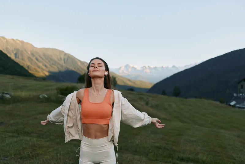 A joyful woman stands in a lush meadow with arms outstretched, eyes closed, embracing nature. She wears a coral sports bra, light jacket, and pale shorts, radiating energy, serenity, and freedom in