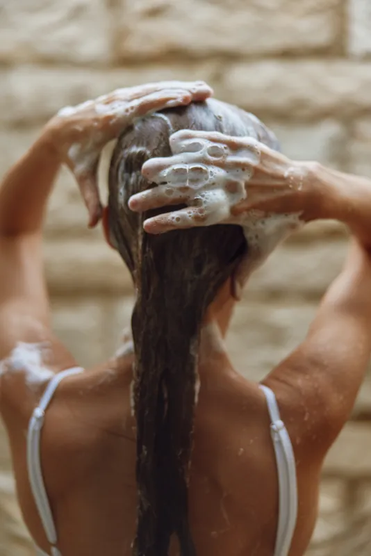 Woman washing hair with foamy shampoo, wet strands covered in bubbles, cleansing routine under warm lighting, natural care and hygiene focus.