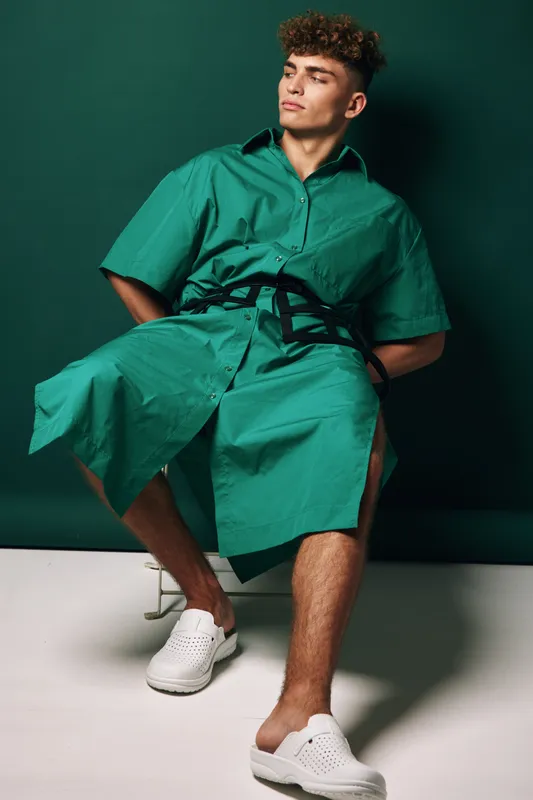 Young man with curly hair wearing green oversized shirt dress and white slip-on shoes sitting on chair against dark green background in studio fashion portrait.