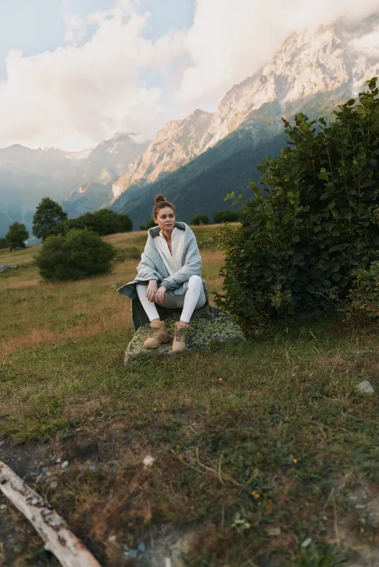 A woman sits on a rocky outcrop in a field with distant mountains, wearing casual outdoor attire. A calm lifestyle moment that highlights nature, serenity, and reflective mood.