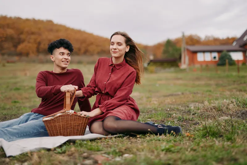 Young couple sharing a candid outdoor picnic in an autumn meadow, genuine connection and natural interaction captured with warm tones, simple props and believable everyday style.