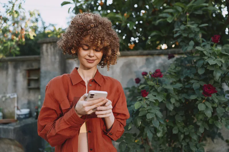 Young woman with curly hair gazes at her smartphone in a sunlit garden, wearing a rust colored shirt and a relaxed casual vibe