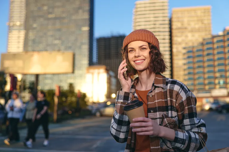 Woman with coffee and phone in city at golden hour, smiling candidly in a beanie, authenticity and mindful living captured in emotional storytelling and urban lifestyle glow.