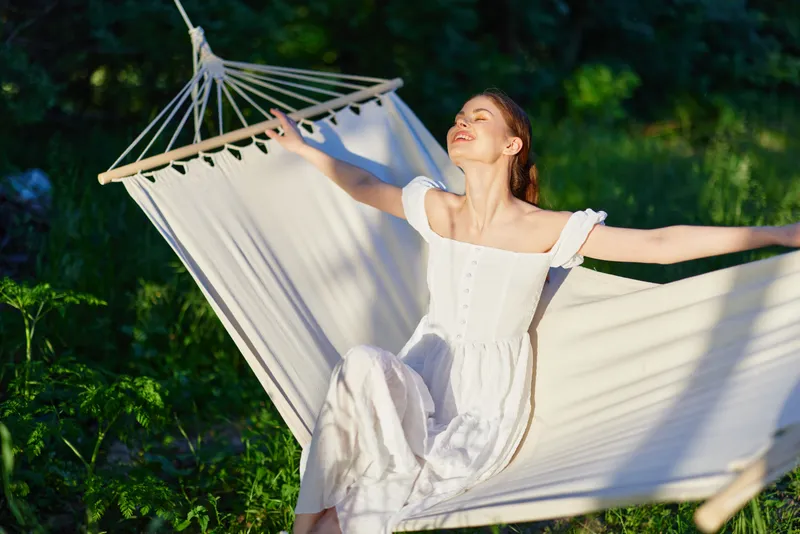 Young woman in a white dress enjoying relaxation in a hammock outdoors, surrounded by greenery, conveying tranquility and blissful summer vibes