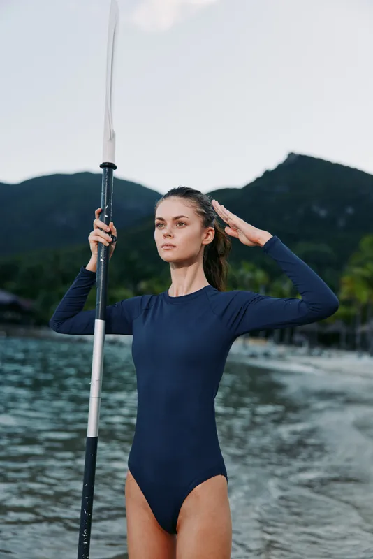 Athletic woman stands in calm water wearing a navy one piece, gripping a spear upright while gazing into the distance; rugged mountains rise behind for a scene of strength and focus.