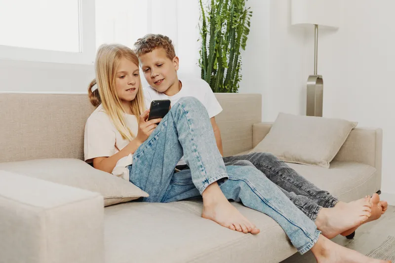 Two barefoot kids sitting on a beige sofa using a smartphone together in a bright modern living room with plants and natural light. Concept of technology and childhood interaction.